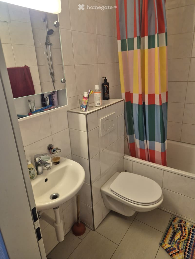 Modern bathroom with a sink, toilet, shower, and mirror. Features a colorful shower curtain and a towel on the floor.