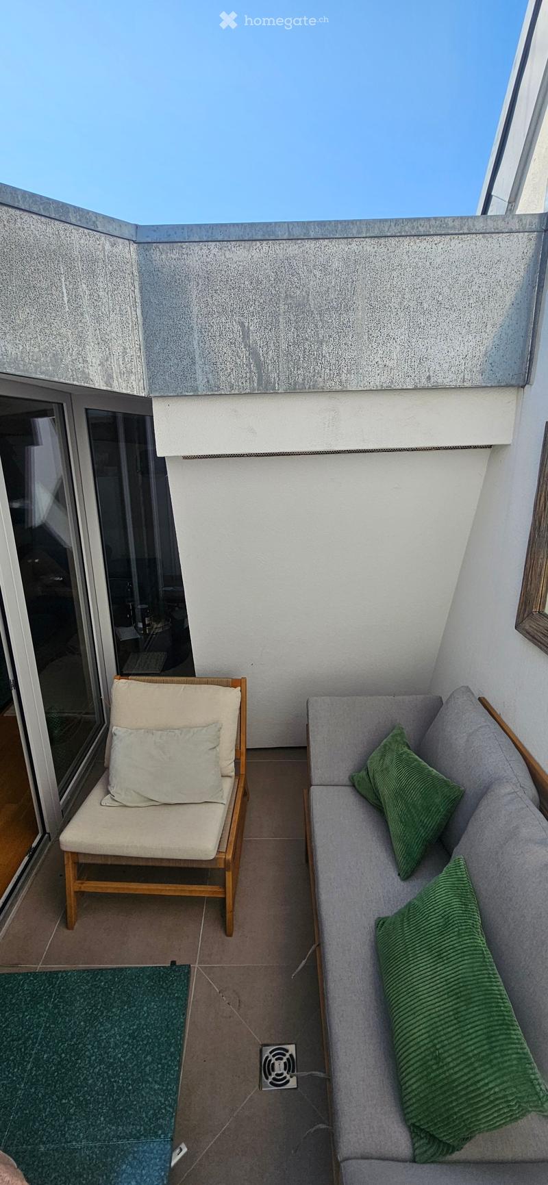 Balcony with a grey sofa, wooden chair, green cushions, and a small green rug.