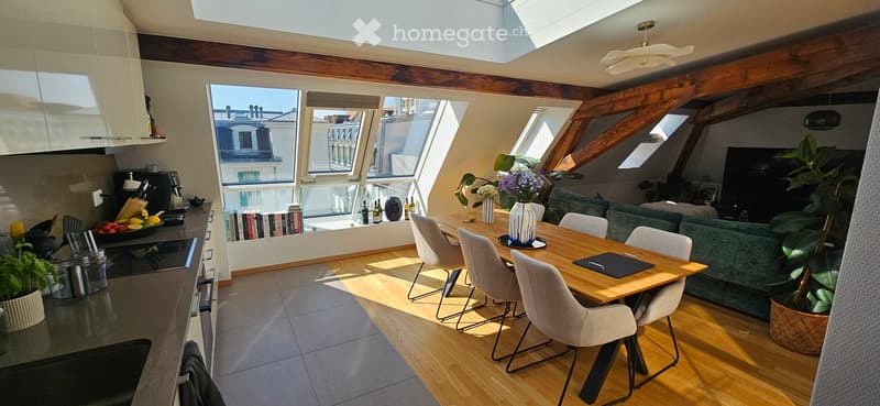 Bright, open-plan living and dining area with wooden floors, large windows, and skylights. Features a green sofa, wooden dining table with chairs, kitchen area with appliances, and decorative plants.