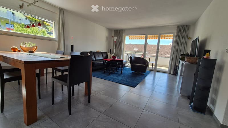Bright, modern living and dining area with large windows and sliding glass doors leading to a balcony. Features a wooden dining table with chairs, a black leather sofa, an armchair, a small side table, a television, and decorative plants.
