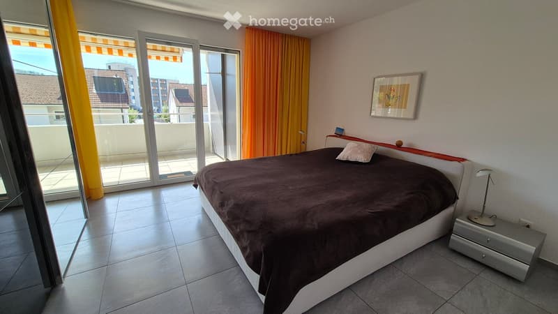 Modern bedroom with a large bed, nightstand, lamp, and picture frame. Large sliding glass doors open to a balcony with a view of the city.