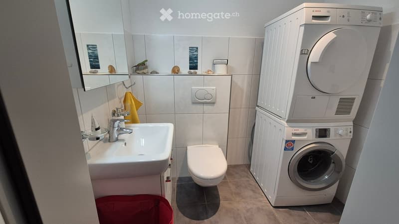 Modern bathroom with a white sink, mirror, toilet, and stacked Bosch washing machine and dryer. A yellow towel hangs on a rack, and decorative items are placed on the mirror shelf.