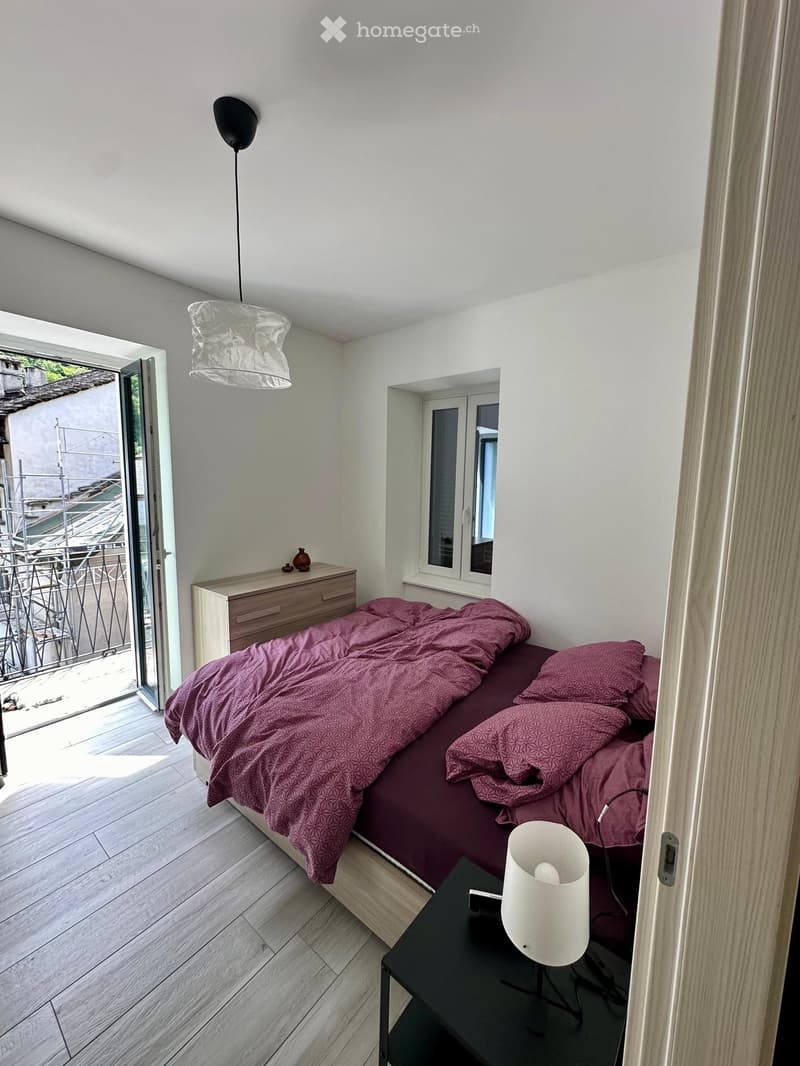 Modern bedroom with a wooden bed, purple bedding, a nightstand with a lamp, a small drawer unit, and a vase on top.