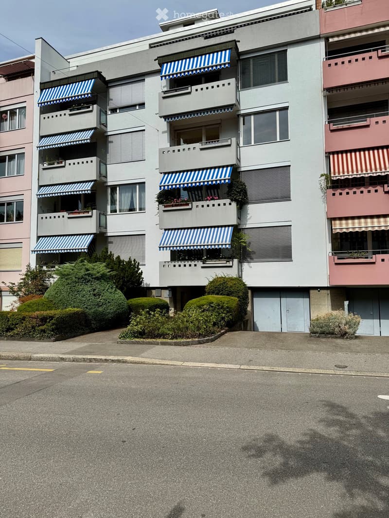 Multi-story residential building with balconies, striped awnings, and surrounding greenery.