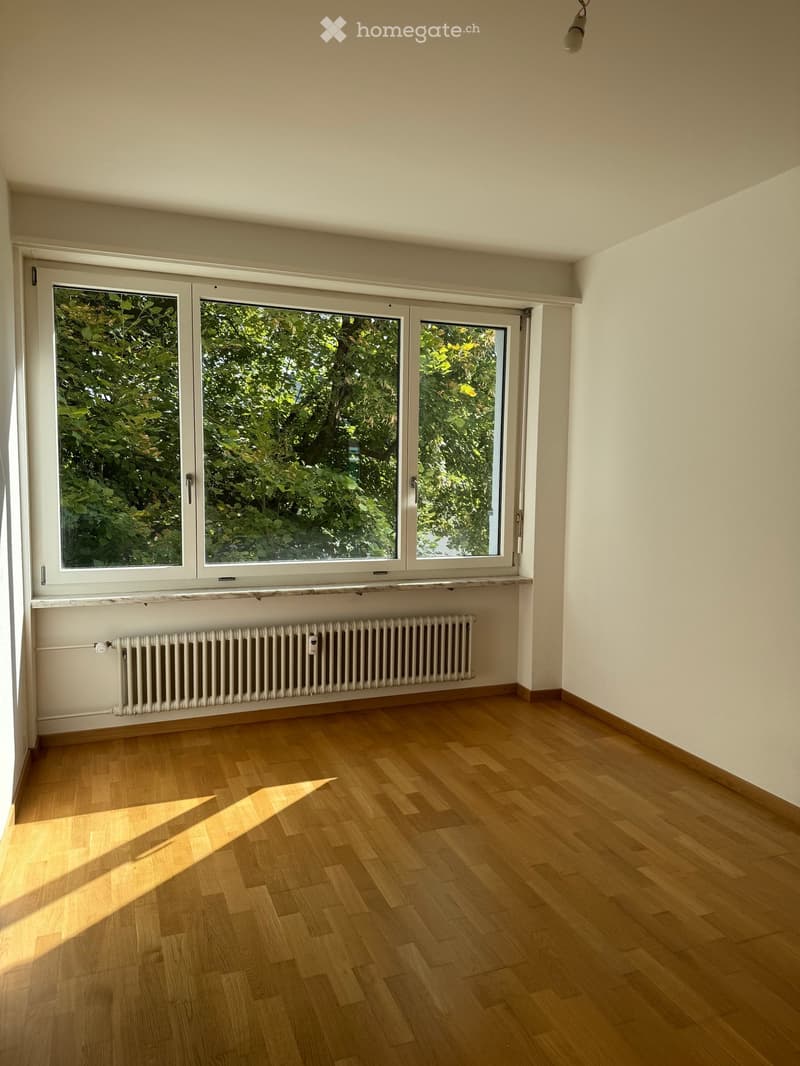 Empty room with wooden flooring, large windows, radiator, and natural light from outside.