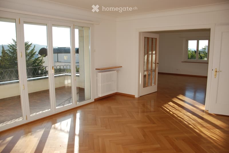 Empty room with wooden flooring, white walls, large sliding glass doors leading to a balcony, and a radiator. Sunlight streams in from the balcony area.