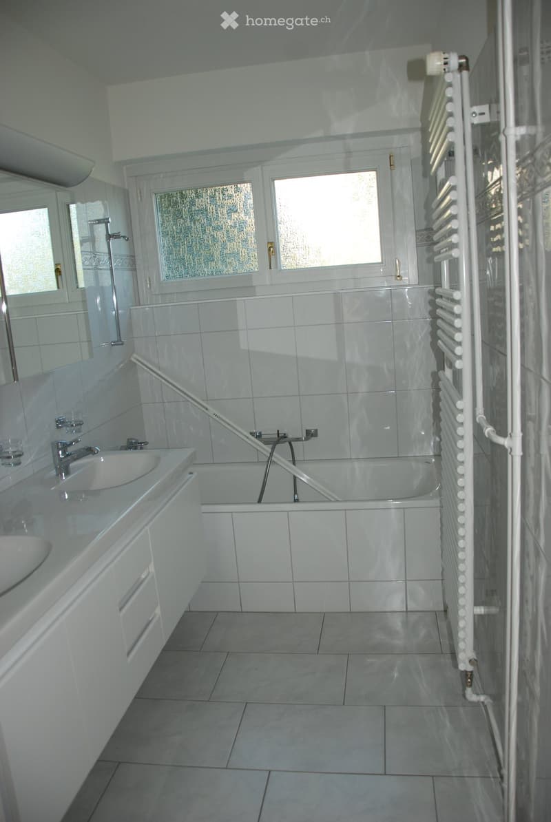 Modern bathroom with white tiles, a double sink, bathtub, radiator, and large windows with frosted glass.