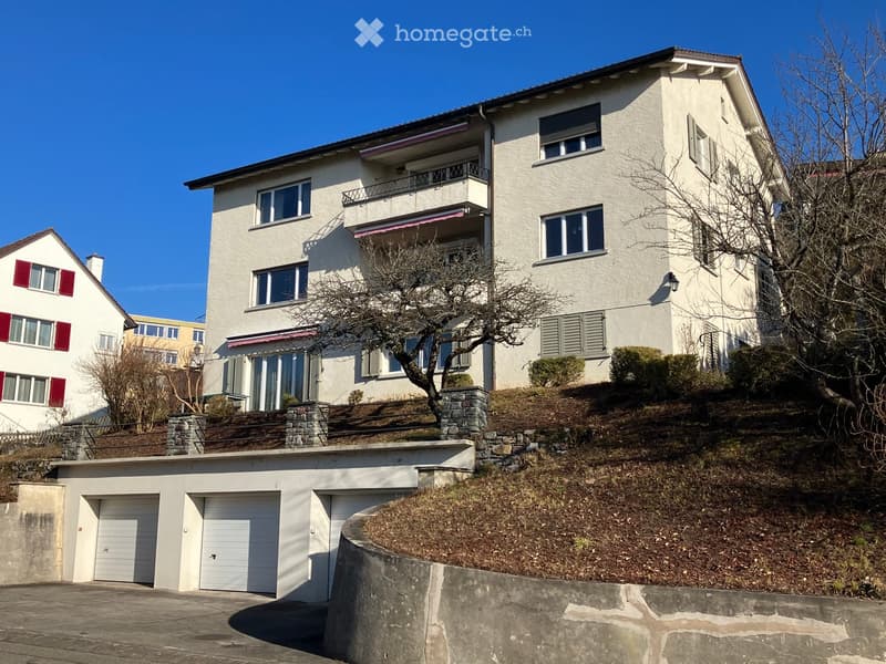 A multi-story white house with balconies, large windows, and a garage. The house is situated on a hillside with a stone retaining wall and a driveway leading to the garage.