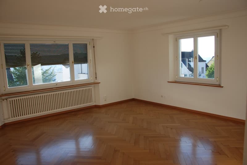 Empty room with wooden flooring, white walls, large windows with blinds, and radiators beneath the windows.