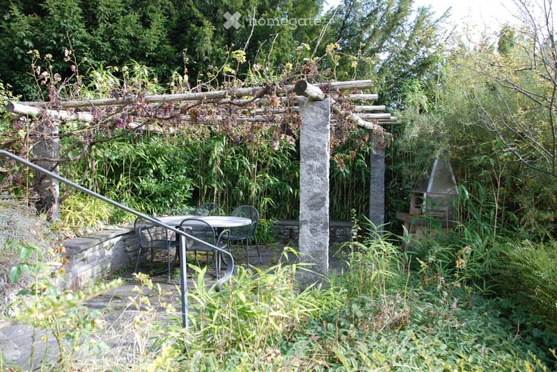 A lush garden area with a stone pergola covered in vines, a round metal table with four chairs, and a brick barbecue surrounded by dense greenery and plants.