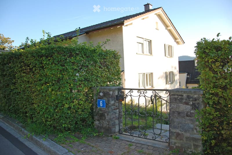 A two-story house with white walls and multiple windows, partially obscured by greenery and a stone gate with the number 6.