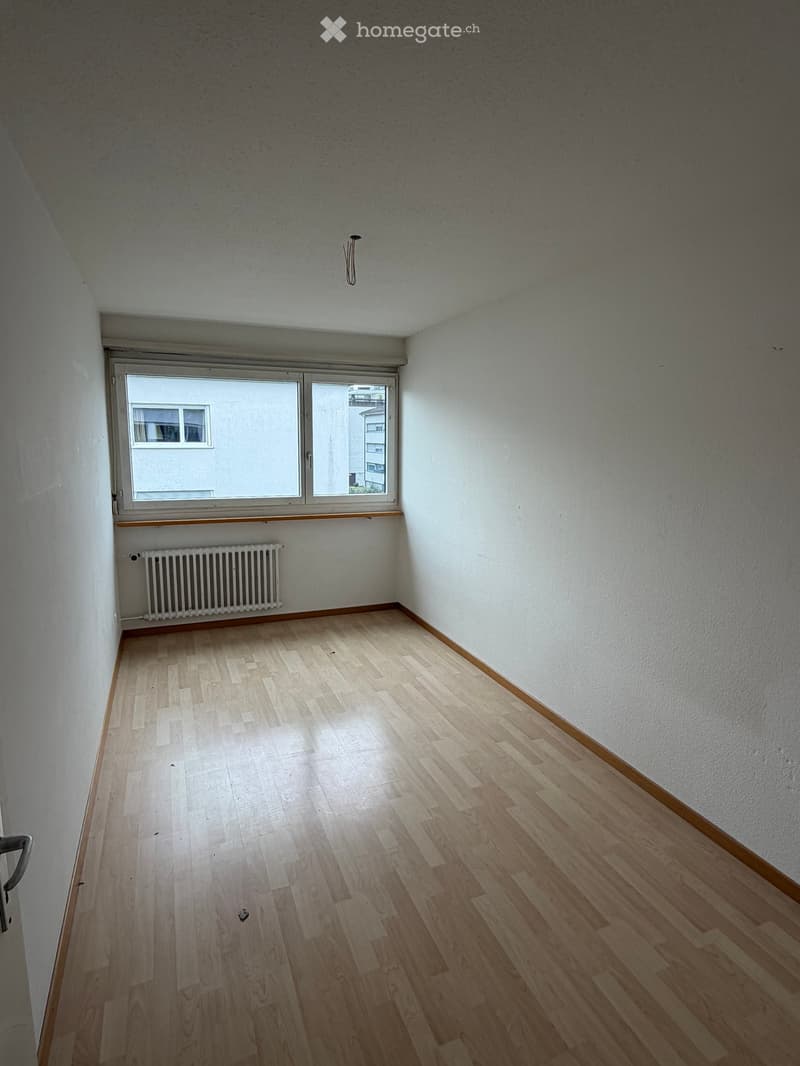 Empty room with light wooden flooring, white walls, and a window with a radiator below it.