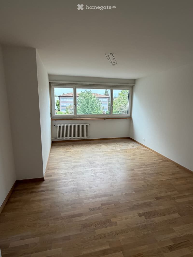 Empty room with wooden flooring, white walls, large window with view, radiator, and visible electrical outlet.