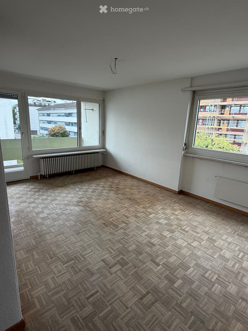 Empty room with wooden herringbone flooring, white walls, large windows, and radiators.
