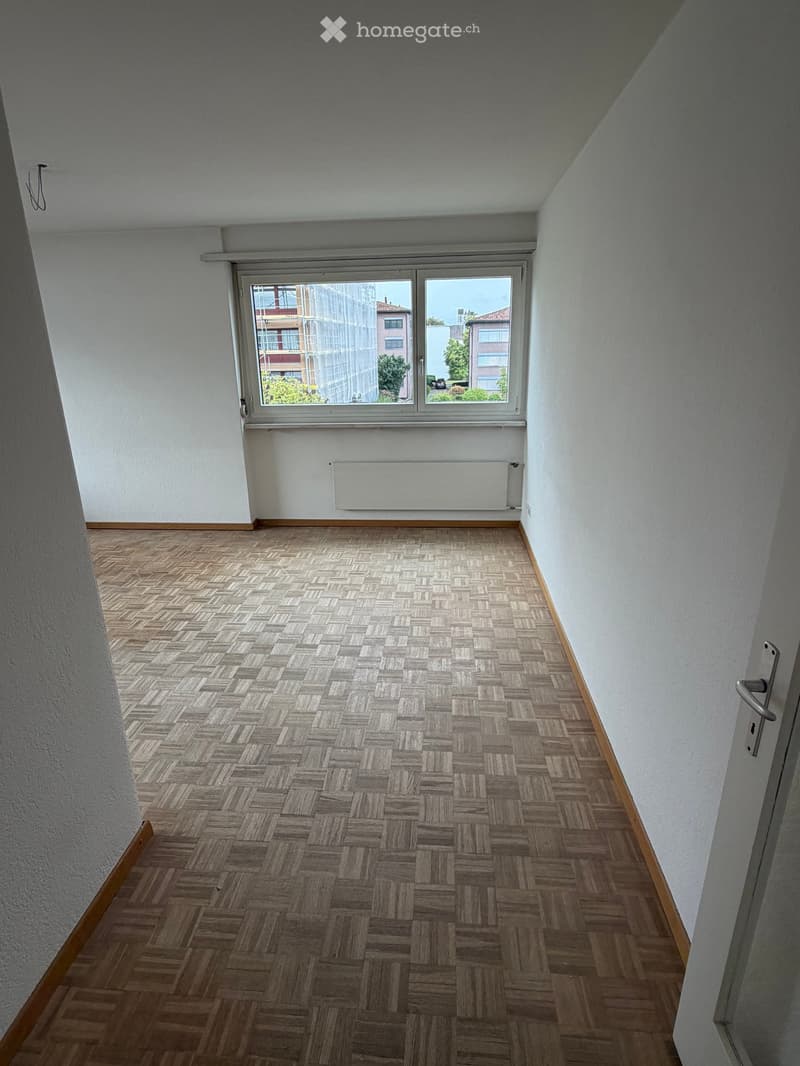 Empty room with wooden herringbone flooring, white walls, and a large window with a view of the city.