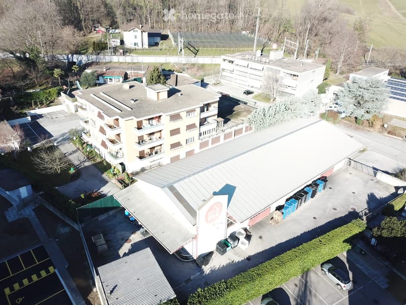 Aerial view of a multi-story residential building with balconies, surrounded by other buildings, parking areas, and greenery.