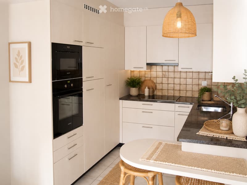 Modern kitchen with white cabinets, a built-in oven, a round dining table with two stools, a sink with a faucet, and decorative items including potted plants, a vase, and a picture frame.