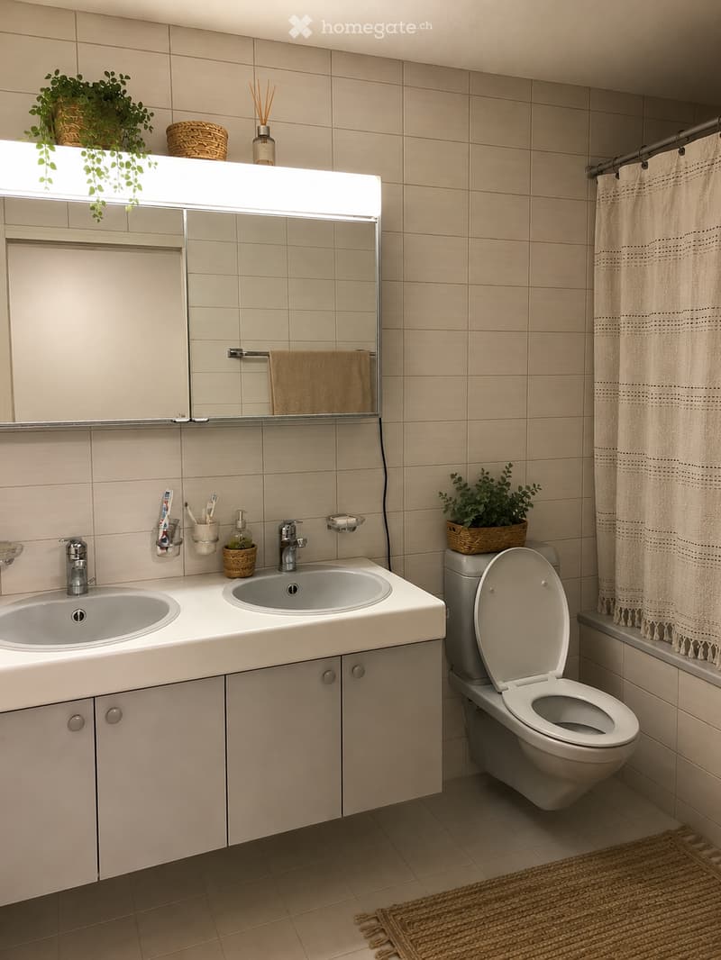 Modern bathroom with beige tiles, double sink, toilet, mirror with built-in lighting, and decorative plants on the windowsill.