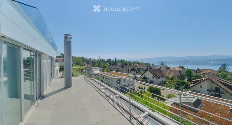 Balcony with modern glass railing, overlooking a scenic town and lake with distant mountains.