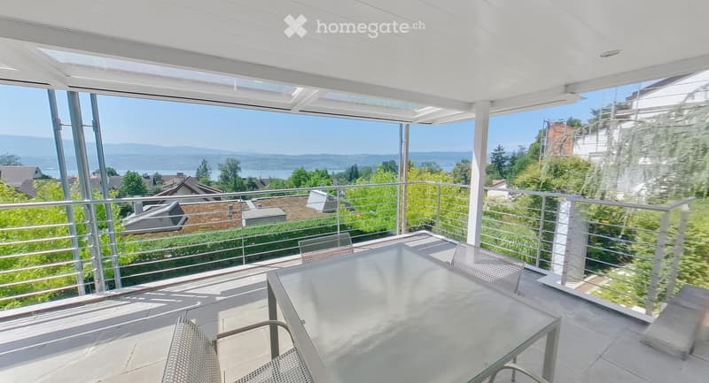 Modern balcony with a glass table and chairs, offering a scenic view of a lake and surrounding landscape.