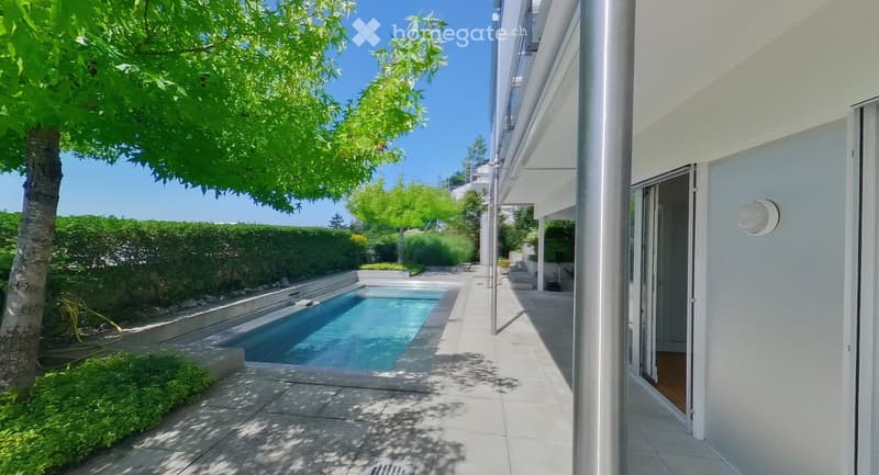 Modern outdoor area with a swimming pool, surrounded by greenery and a well-maintained garden. The area features a paved walkway and a white building with large windows in the background.