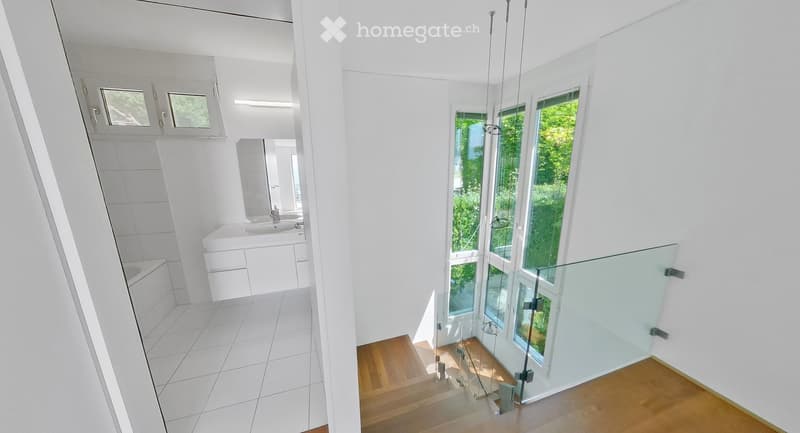 Modern, minimalist hallway with white walls, wooden flooring, and large windows offering a view of greenery. A glass railing is visible on the staircase leading upwards.
