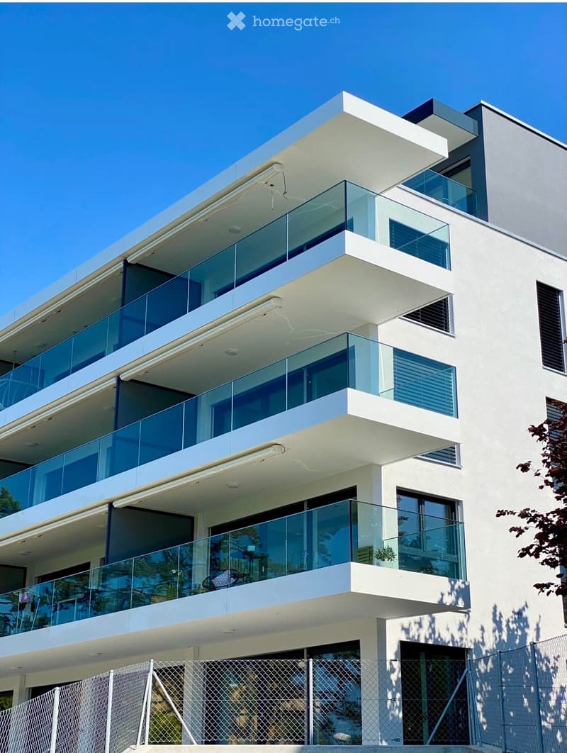 Modern multi-story building with white facade, large glass windows, and balconies with glass railings.