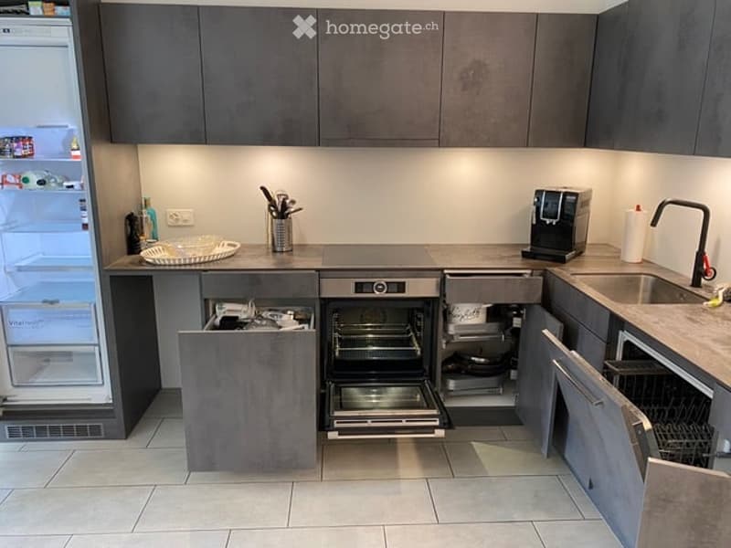 Modern kitchen with grey cabinets, stainless steel appliances, and a clean, organized layout. Features include a refrigerator, oven, dishwasher, sink, and coffee maker.