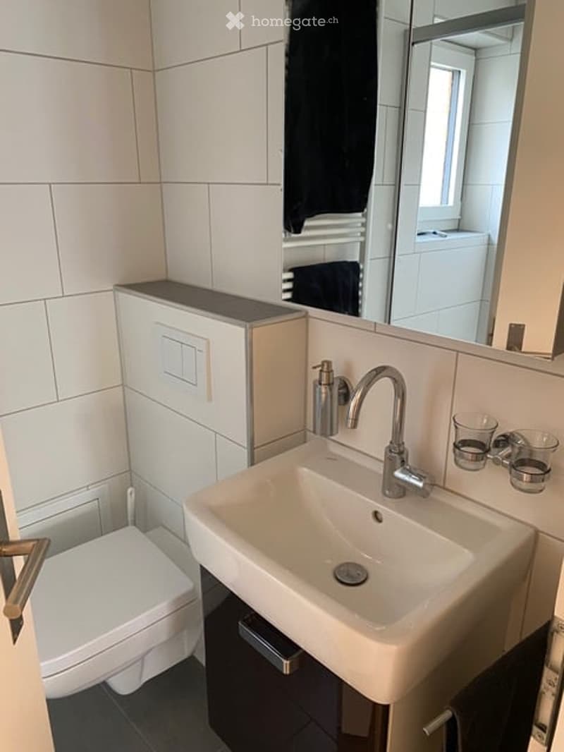 Modern bathroom with white tiles, a sink with a chrome faucet, a toilet, a large mirror, and two small cups on the sink counter.