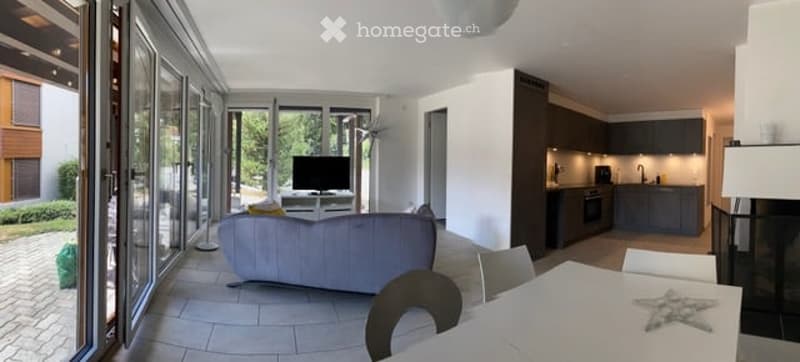 Bright, open-plan living and dining area with large glass doors leading to a garden view. Features a grey sofa, coffee table, TV, dining table with chairs, and a modern kitchen with cabinets, oven, and sink.