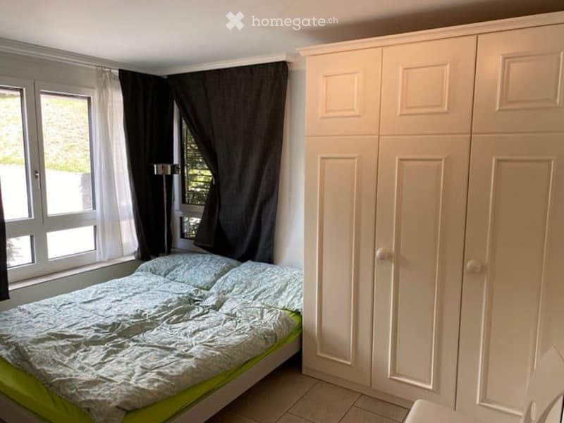 A tidy bedroom with a double bed, white wardrobe, and large windows with dark curtains.