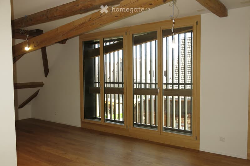 Empty room with wooden flooring, exposed wooden beams on the ceiling, and large wooden-framed windows with metal bars.