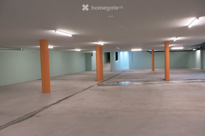 Empty underground parking area with concrete floors, light blue walls, and orange support pillars. The space is well-lit with fluorescent lighting and has a clean, unused appearance.