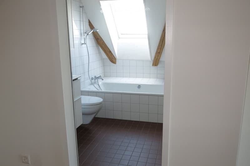Bathroom with a bathtub, toilet, and a window above the tub. The walls are tiled, and the floor is covered with dark tiles.