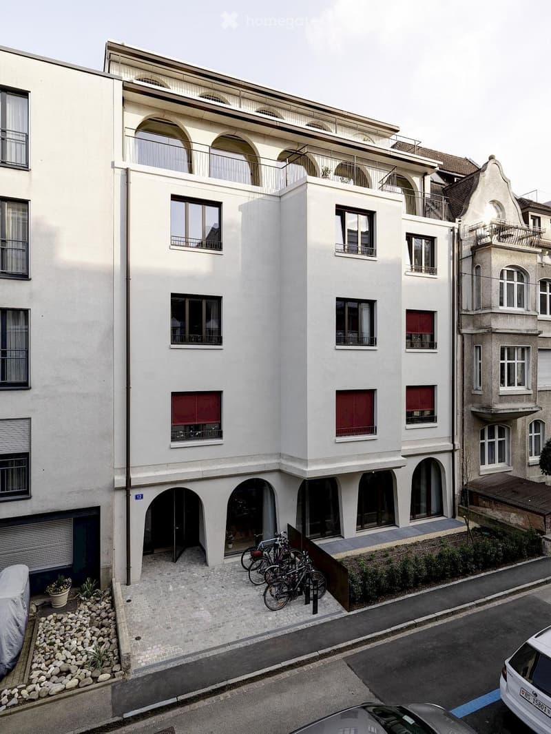 Modern multi-story building with white facade, arched windows, and balconies. Bicycles parked in front, small garden area with potted plants, and a covered car parked nearby.