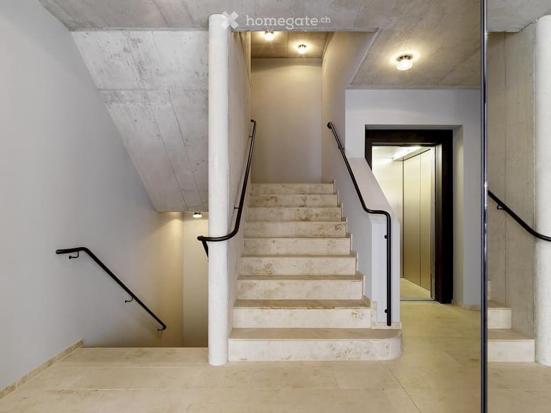 Modern interior staircase with beige stone steps, black metal railings, and recessed lighting, leading to an open area with white walls and a raised ground floor.