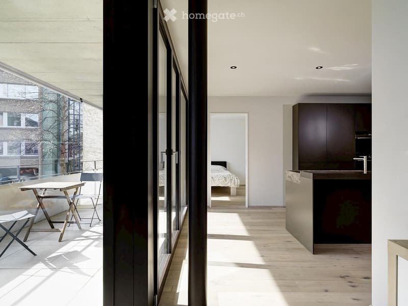 Modern, open-plan living space with a kitchen area, a bedroom visible through a doorway, and a small outdoor seating area with a table and chairs.