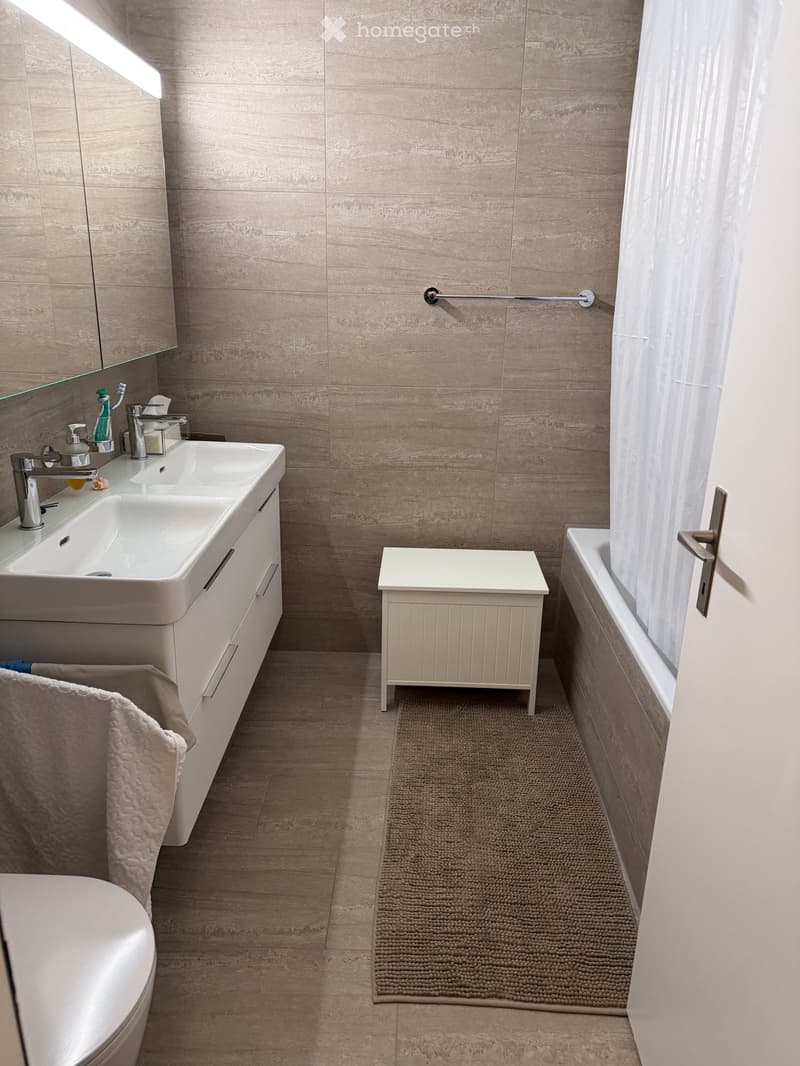 Modern bathroom with beige tiles, white fixtures, double vanity with two sinks, bathtub, shower with white curtain, wooden stool, small rug, white cabinet, and mirror.