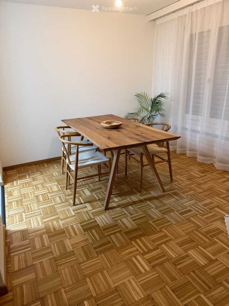 dining area with a wooden table and four chairs, a bowl on the table, white curtains, wooden parquet floor