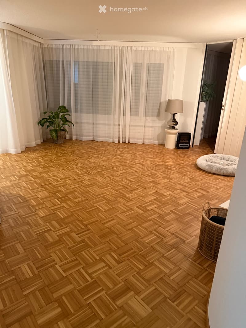 Empty room with wooden parquet flooring, white curtains, a white lamp on a small pedestal, and some potted plants.