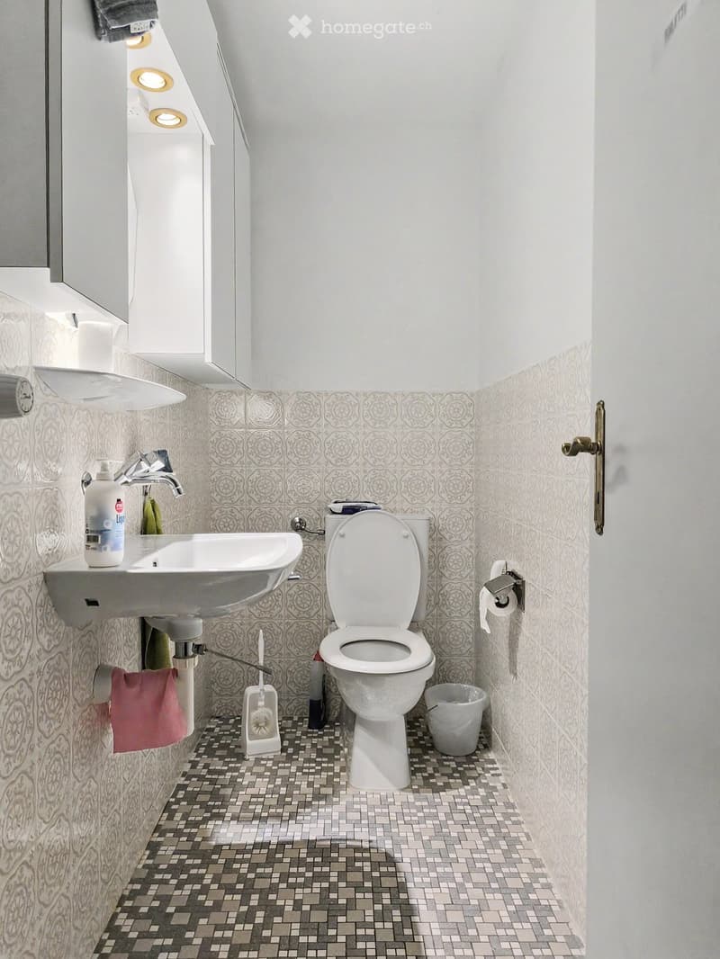 Modern bathroom with white walls, patterned tiles, a toilet, sink, and a small trash can. Includes a towel and a bottle on the sink.