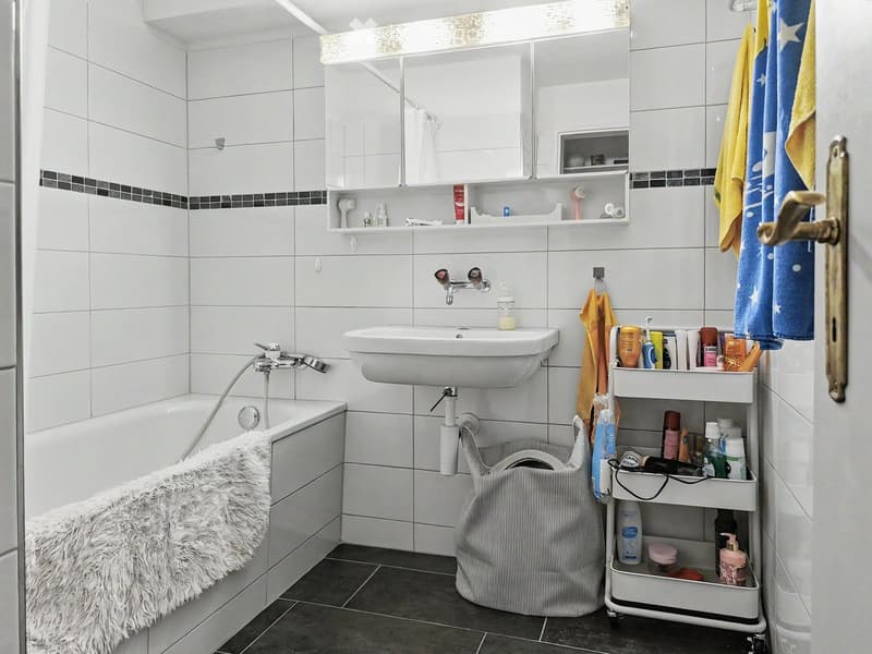 Modern bathroom with white tiles, bathtub, sink, mirror, and a cart holding toiletries.