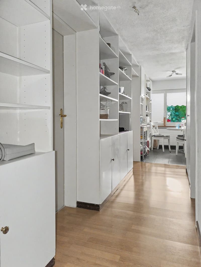 A modern, minimalist hallway with white walls, light wooden flooring, built-in white shelving units, and a small adjacent room with a table and chairs.