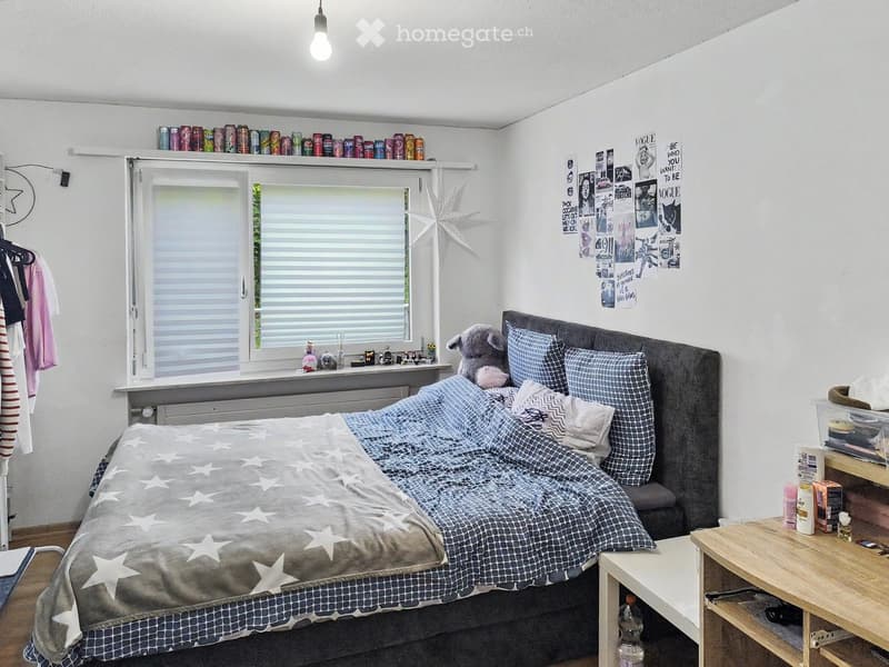 A cozy bedroom with a large bed, nightstand, and a window with blinds. The room features posters on the wall, a shelf with cans, and a small table with various items.