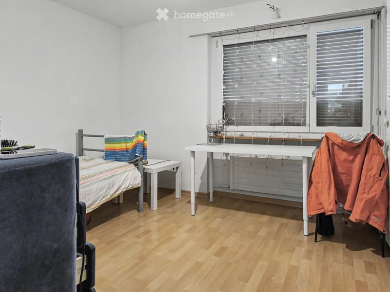 A small, well-lit bedroom with a bed, desk, and chair. The room features wooden flooring and white walls, with a window covered by blinds and string lights.