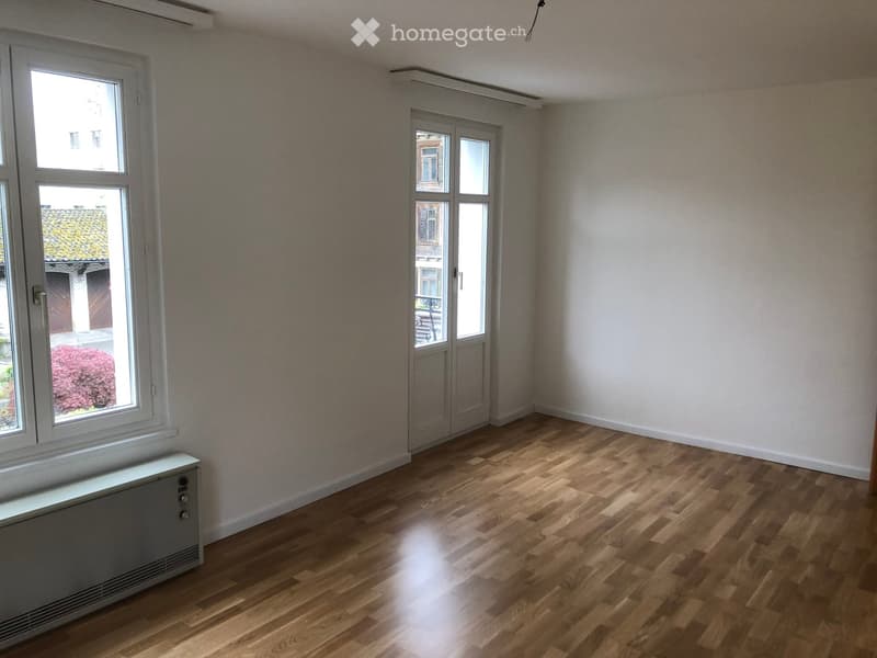 Empty room with wooden flooring, large windows, and a radiator. No furniture or equipment present.