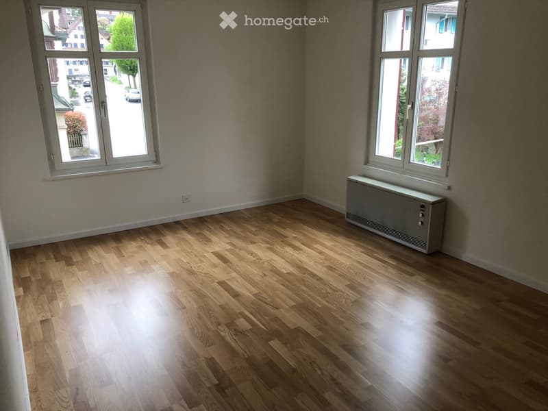 Empty room with wooden flooring, large windows, radiator, and neutral-colored walls.