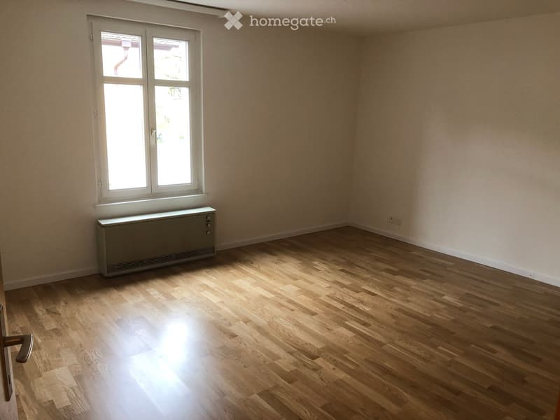 Empty room with wooden flooring, light-colored walls, and a window with white frames. A radiator is placed below the window.
