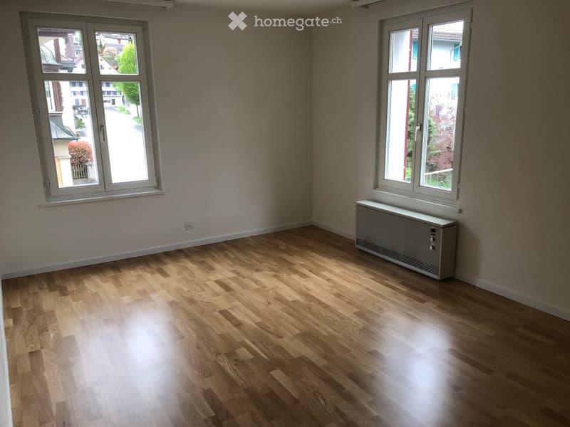 Empty room with wooden flooring, large windows, radiator, and neutral-colored walls.