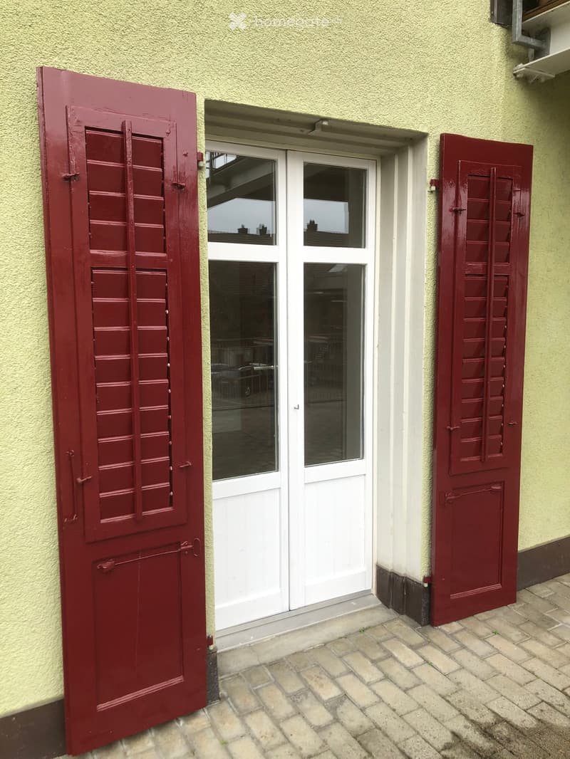 Exterior view of a building entrance with a white double door flanked by red shutters, set in a light yellow wall with a brick-paved ground.